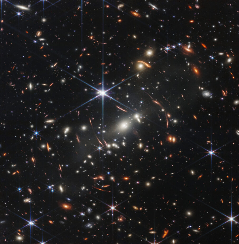 Φωτογραφία της ομάδας γαλαξιών SMACS 0723, η πιο λεπτομερής φωτογραφία πολύ μακρινών γαλαξιών που τραβήχτηκε ποτέ στο υπέρυθρο. Καλύπτει τμήμα του ουρανού, όσο ένας κόκκος άμμου σε απόσταση απλωμένου χεριού. Προέκυψε από τη σύνθεση πολλών φωτογραφιών με έκθεση 2 ωρών η καθεμιά, ώστε να συλλάβουν αρκετά φωτόνια από τα αμυδρά ουράνια σώματα που απεικονίζονται. Το σώμα με τις ακτίνες είναι άστρο και οι ακτίνες τεχνούργημα των κατόπτρων